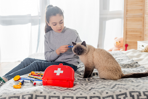 girl making injection to cat with toy syringe near first aid kit on bed