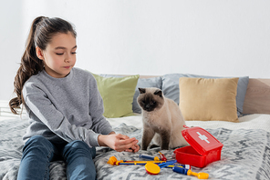 girl sitting on bed and playing doctor with toy medical set near cat