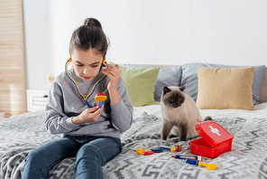 girl with toy stethoscope playing doctor on bed near cat and first aid kit