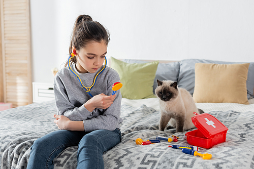 girl holding toy stethoscope while playing on bed near cat