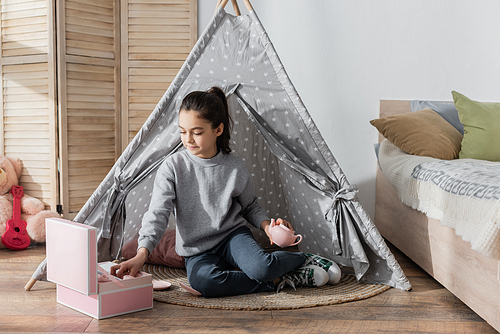 girl holding teapot while sitting in wigwam near box with toy tea set