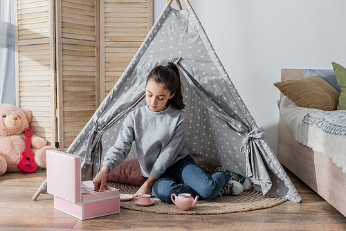 preteen girl playing with toy tea set while sitting on floor in wigwam