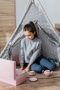 brunette preteen girl sitting on floor in wigwam and playing with toy tea set