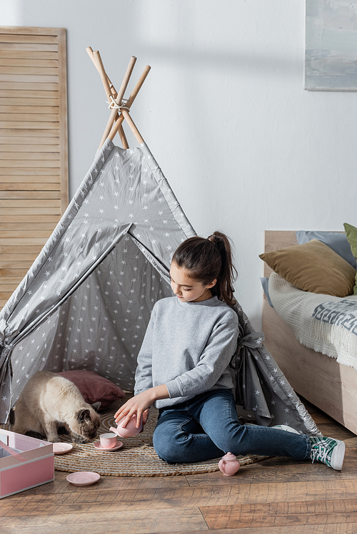 girl pouring tea from toy teapot while playing with cat in wigwam