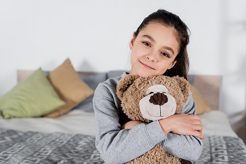 cheerful preteen girl embracing teddy bear and  in bedroom