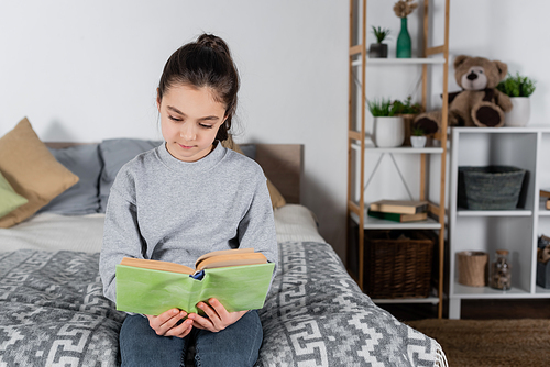 brunette preteen girl sitting on bed and reading book