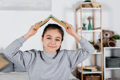 cheerful kid holding book above head and smiling at camera