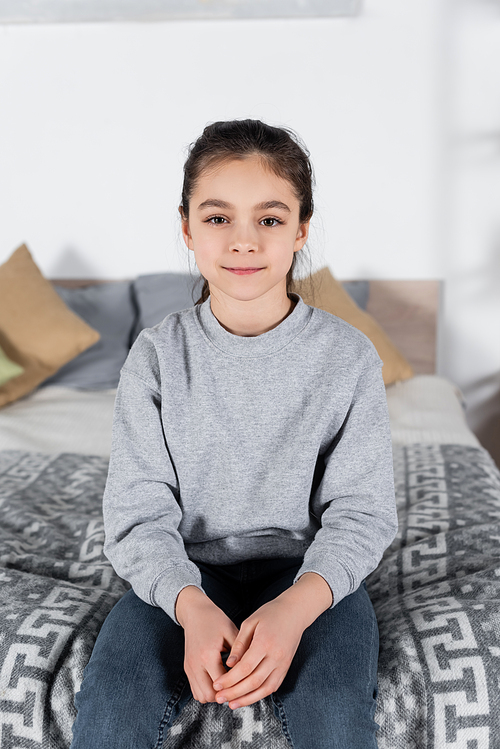 preteen girl smiling at camera while sitting on blurred bed