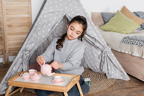 preteen girl sitting in wigwam and pouring tea from toy teapot