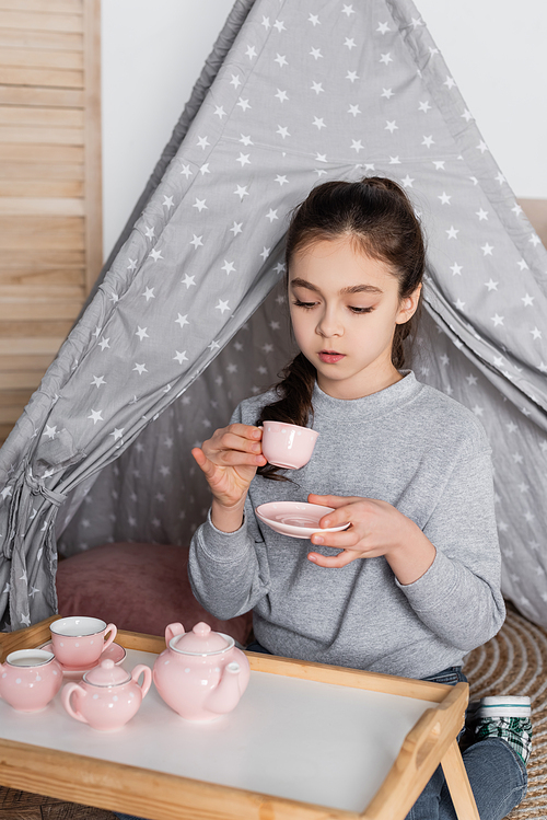 girl drinking tea from toy cup while playing in wigwam