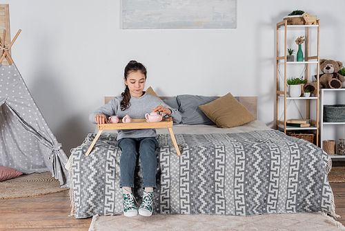 full length view of girl sitting on bed with toy tea set on wooden tray