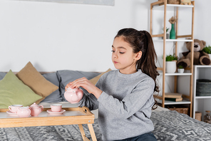 girl pouring tea from toy teapot while sitting on bed