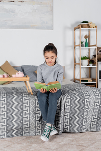 full length view of girl sitting on bed near tray with toy tea set and reading book