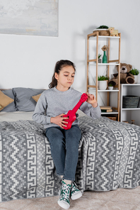 girl tuning toy guitar while sitting on bed at home