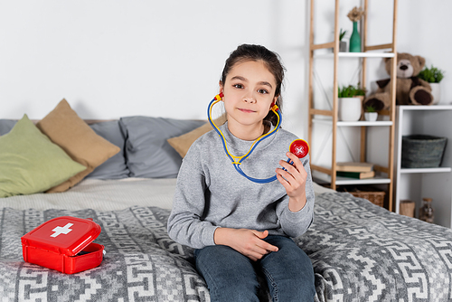girl with toy stethoscope sitting on bed near first aid kit