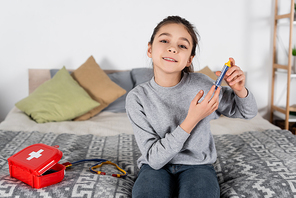 smiling girl holding toy syringe while sitting on bed near first aid kit and stethoscope