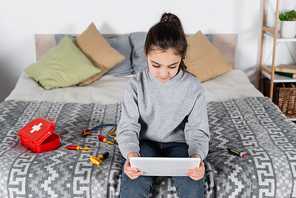 girl using digital tablet while sitting on bed near toy medical set