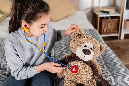 preteen girl examining teddy bear with toy stethoscope while playing at home