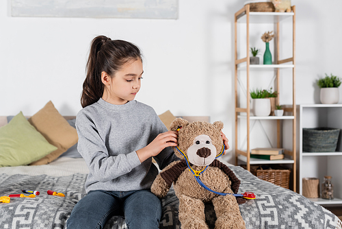girl putting toy stethoscope on teddy bear while playing in bedroom