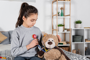 girl playing doctor with teddy bear and toy stethoscope in bedroom