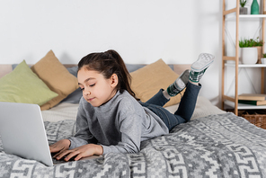 full length view of preteen girl lying on bed and typing on laptop