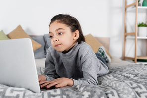 preteen girl lying on bed and typing on laptop