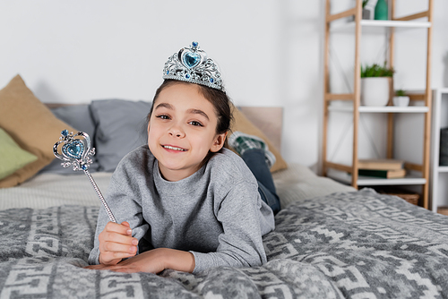 smiling girl in toy crown holding royal wand while lying on bed