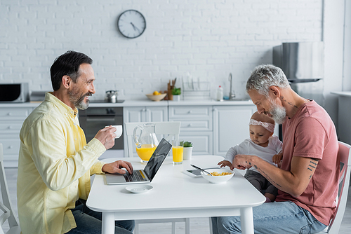 Smiling homosexual man with laptop and coffee looking at partner with daughter in kitchen