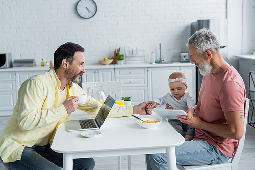 Homosexual parents with devices looking at baby daughter near breakfast in kitchen