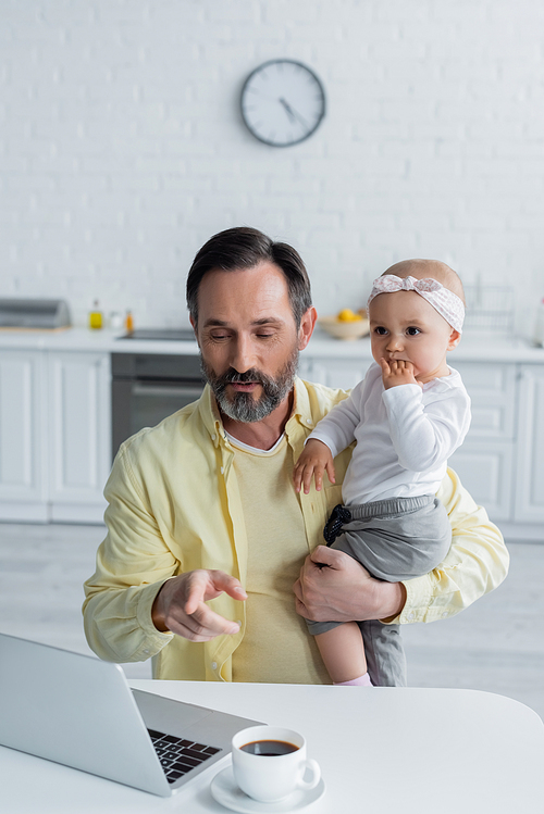 Mature man pointing at laptop while holding baby daughter in kitchen