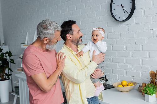 Cheerful homosexual parents holding child in kitchen