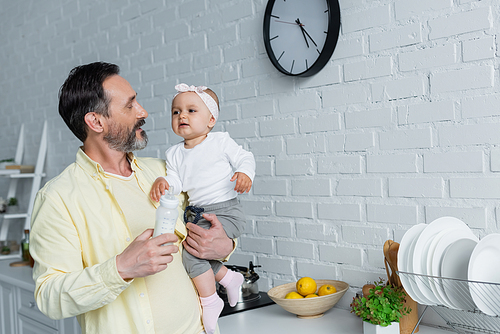 Mature man holding baby daughter and bottle of milk in kitchen