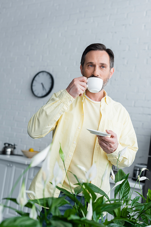 Mature man drinking coffee near plant in kitchen