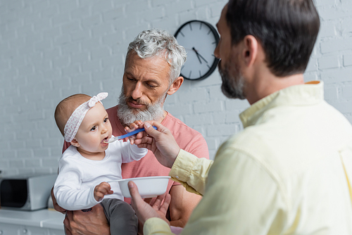 Homosexual man holding baby daughter near partner with spoon and plate