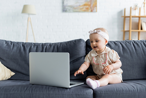 Cheerful baby girl sitting near laptop on couch