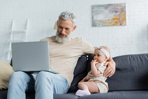 Mature man holding laptop and embracing baby girl on couch