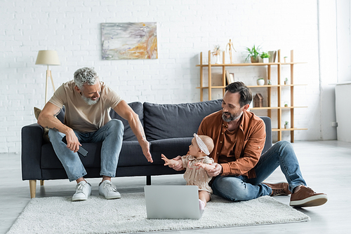 Smiling homosexual couple playing with baby daughter near laptop on carpet at home