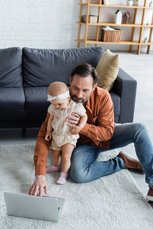 Man using laptop and holding toddler daughter on carpet