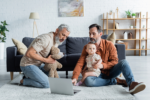 Same sex family with baby daughter using laptop and holding soft toy