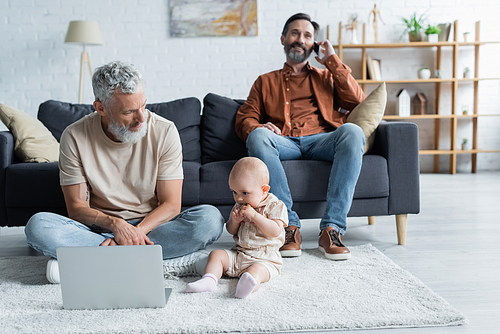 Toddler child sitting near homosexual father with laptop on carpet