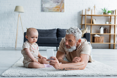 Mature man holding smartphone near toddler daughter at home
