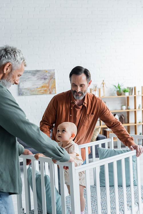 Smiling homosexual parents standing near baby daughter in crib