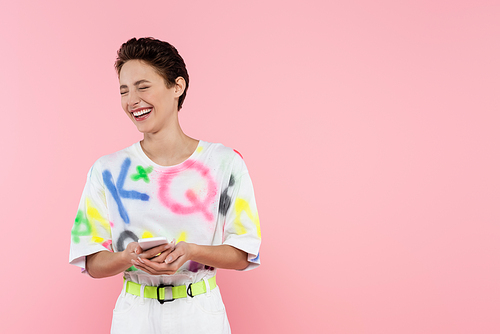 excited woman with closed eyes laughing while holding smartphone isolated on pink