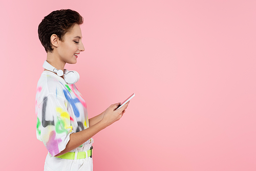 side view of smiling woman with wireless headphones on neck using cellphone isolated on pink