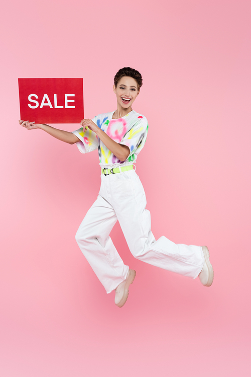 excited woman in stylish clothes jumping with red card with sale lettering on pink