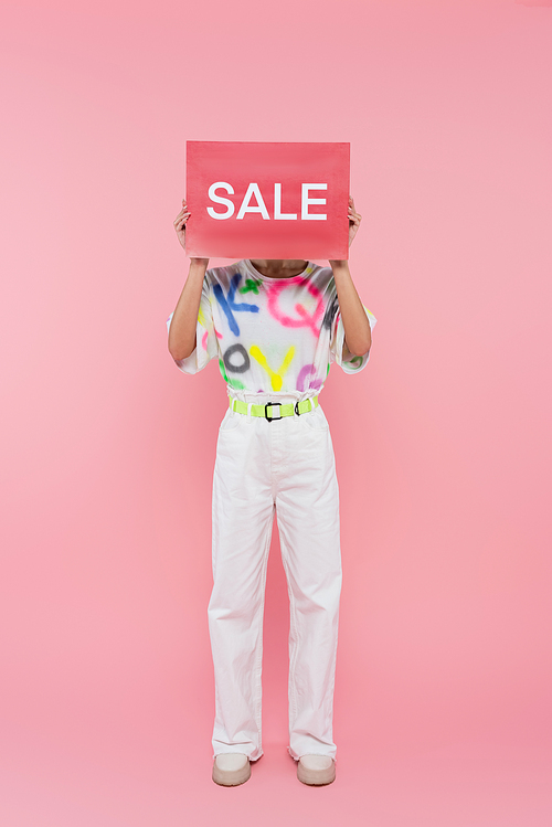 full length view of woman in white pants hiding face behind card with sale lettering on pink