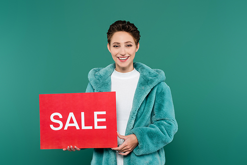 happy woman in faux fur jacket  while holding card with sale lettering isolated on green