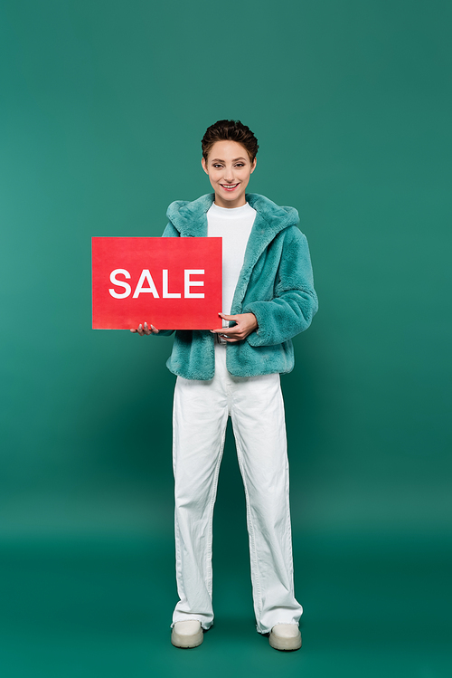 full length view of happy woman in white pants and faux fur jacket holding red card with sale lettering on green