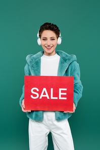 brunette woman in faux fur jacket and headphones holding red card with sale lettering isolated on green