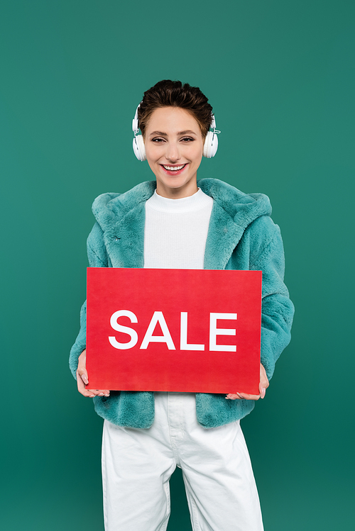 brunette woman in faux fur jacket and headphones holding red card with sale lettering isolated on green
